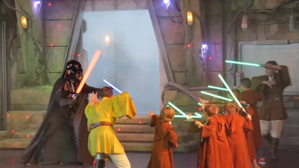 Des enfants habillés en stagiaires de l'Académie de formation Jedi tenant des sabres laser affrontent Dark Vador lors d'un événement sur le thème de Star Wars avec un fond enfumé et un éclairage théâtral.