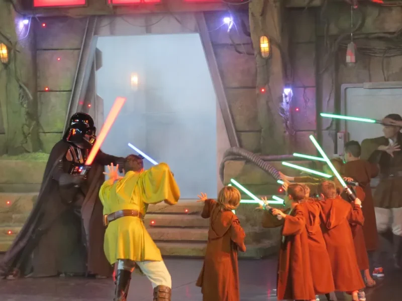 Des enfants habillés en stagiaires de l'Académie de formation Jedi tenant des sabres laser affrontent Dark Vador lors d'un événement sur le thème de Star Wars avec un fond enfumé et un éclairage théâtral.