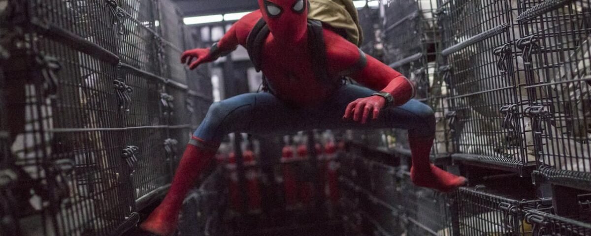 Spider-man, dans une scène de "Spiderman Homecoming", est accroupi entre d'étroites rangées de cages métalliques, vêtu de son emblématique costume rouge et bleu avec un sac à dos marron, à l'intérieur d'un espace sombre.