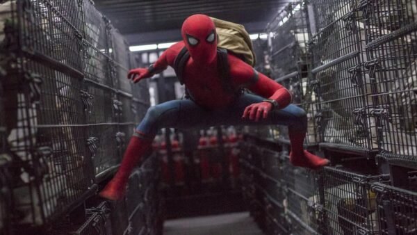 Spider-man, dans une scène de "Spiderman Homecoming", est accroupi entre d'étroites rangées de cages métalliques, vêtu de son emblématique costume rouge et bleu avec un sac à dos marron, à l'intérieur d'un espace sombre.