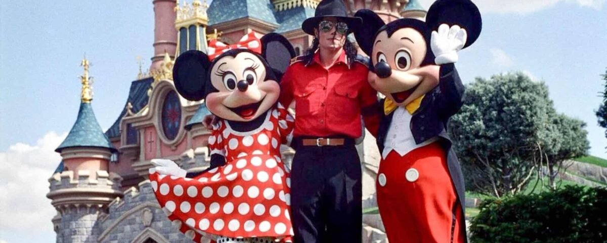 Une personne se tient entre Minnie Mouse et Mickey Mouse devant un château de conte de fées sous un ciel clair et ensoleillé. Minnie porte une robe rouge à pois inspirée de Michael Jackson et Mickey