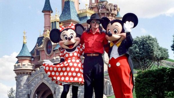Une personne se tient entre Minnie Mouse et Mickey Mouse devant un château de conte de fées sous un ciel clair et ensoleillé. Minnie porte une robe rouge à pois inspirée de Michael Jackson et Mickey