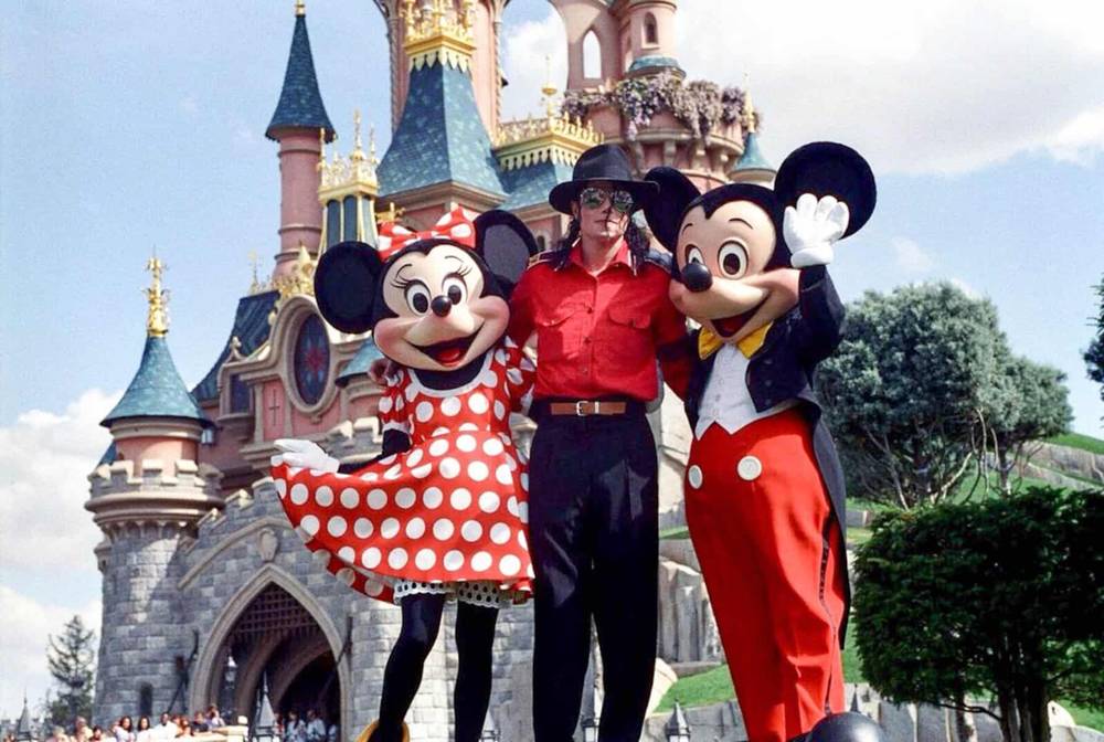 Une personne se tient entre Minnie Mouse et Mickey Mouse devant un château de conte de fées sous un ciel clair et ensoleillé. Minnie porte une robe rouge à pois inspirée de Michael Jackson et Mickey