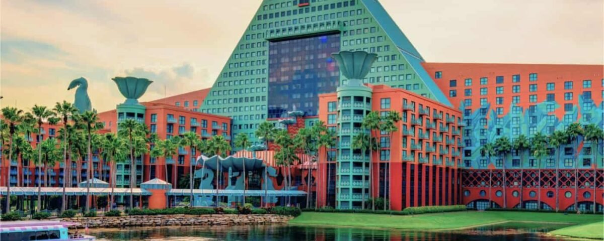 Hôtel Walt Disney World Dolphin en forme de pyramide avec façades vertes et rouge corail au bord d'une rivière paisible, sous un ciel doux, avec un bateau glissant sur l'eau.