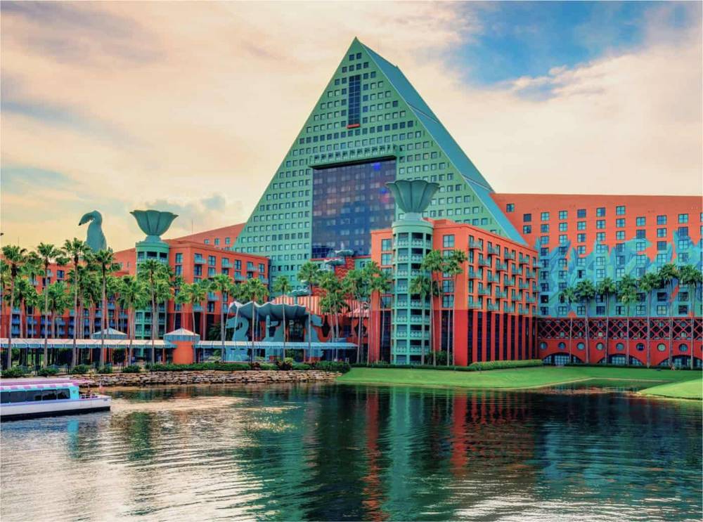 Hôtel Walt Disney World Dolphin en forme de pyramide avec façades vertes et rouge corail au bord d'une rivière paisible, sous un ciel doux, avec un bateau glissant sur l'eau.