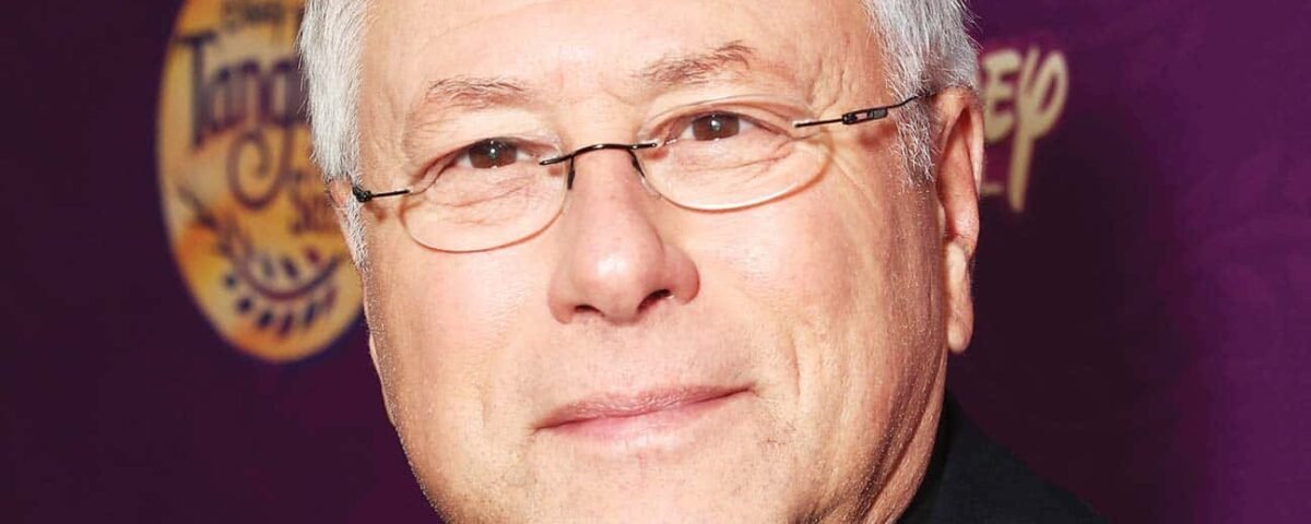 Un homme caucasien plus âgé aux cheveux gris courts et aux lunettes, souriant à la caméra, vêtu d'un costume noir. Il se tient devant un fond violet avec des emblèmes dorés, rappelant Alan Men.