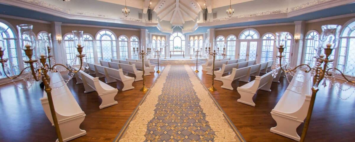 Intérieur élégant de la salle de mariage de conte de fées de Disney avec une longue allée de tapis bleu et or, des chaises blanches alignées de chaque côté, des bougeoirs ornés et de grandes fenêtres sous un plafond voûté avec ch