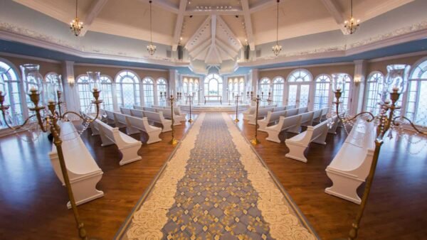 Intérieur élégant de la salle de mariage de conte de fées de Disney avec une longue allée de tapis bleu et or, des chaises blanches alignées de chaque côté, des bougeoirs ornés et de grandes fenêtres sous un plafond voûté avec ch