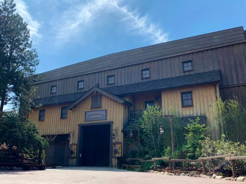 Un grand bâtiment de style grange en bois avec une pancarte indiquant "Théâtre Chaparral" sous un ciel bleu clair, entouré d'arbres verts luxuriants.
