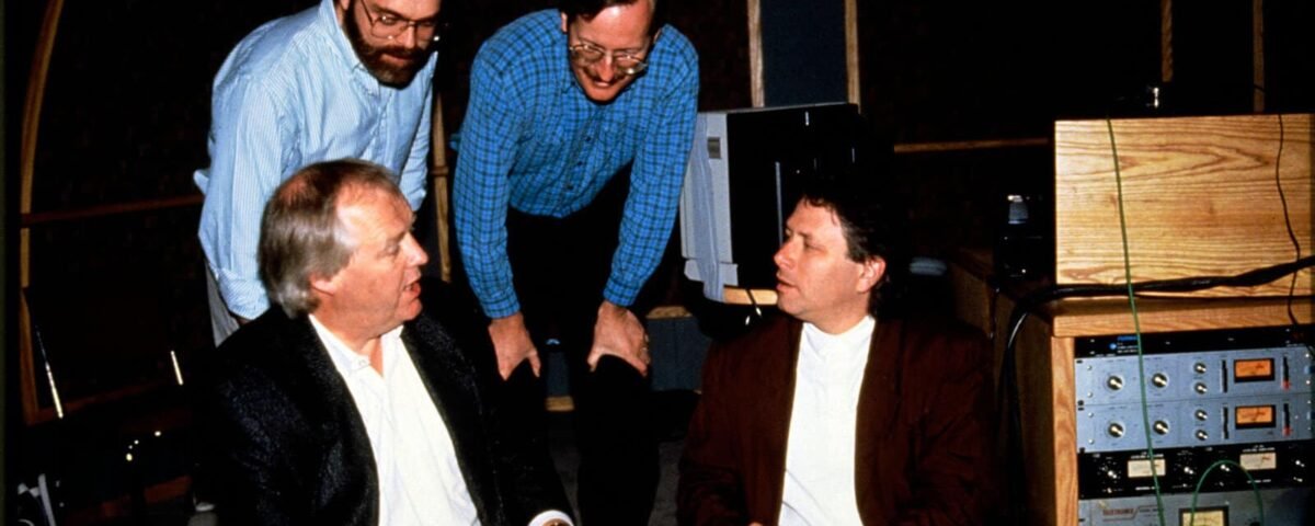 Quatre hommes, un assis et trois debout, engagent une discussion dans un studio d'enregistrement rempli de matériel audio. Alan Menken en fait partie.