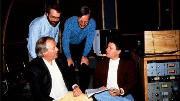 Quatre hommes, un assis et trois debout, engagent une discussion dans un studio d'enregistrement rempli de matériel audio. Alan Menken en fait partie.