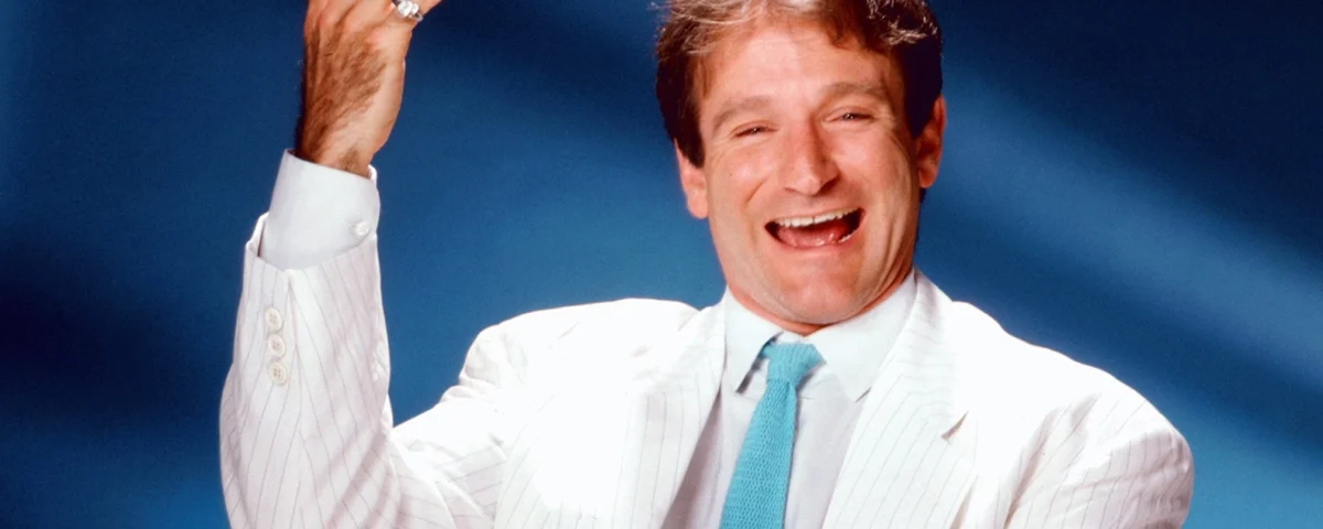 Un joyeux Robin Williams en costume blanc et cravate bleue fait des gestes animés sur un fond bleu. Il semble être en train de rire, levant la main droite dans une pose dynamique.