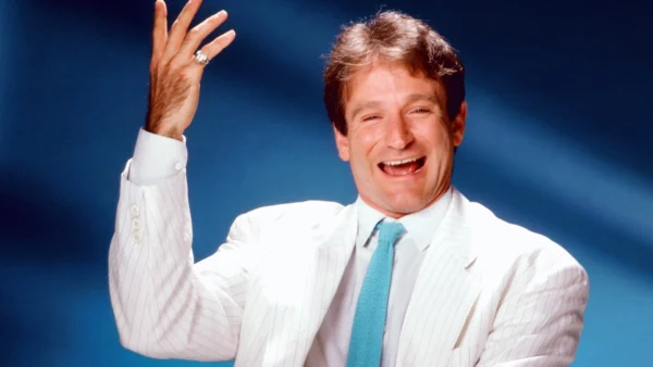 Un joyeux Robin Williams en costume blanc et cravate bleue fait des gestes animés sur un fond bleu. Il semble être en train de rire, levant la main droite dans une pose dynamique.