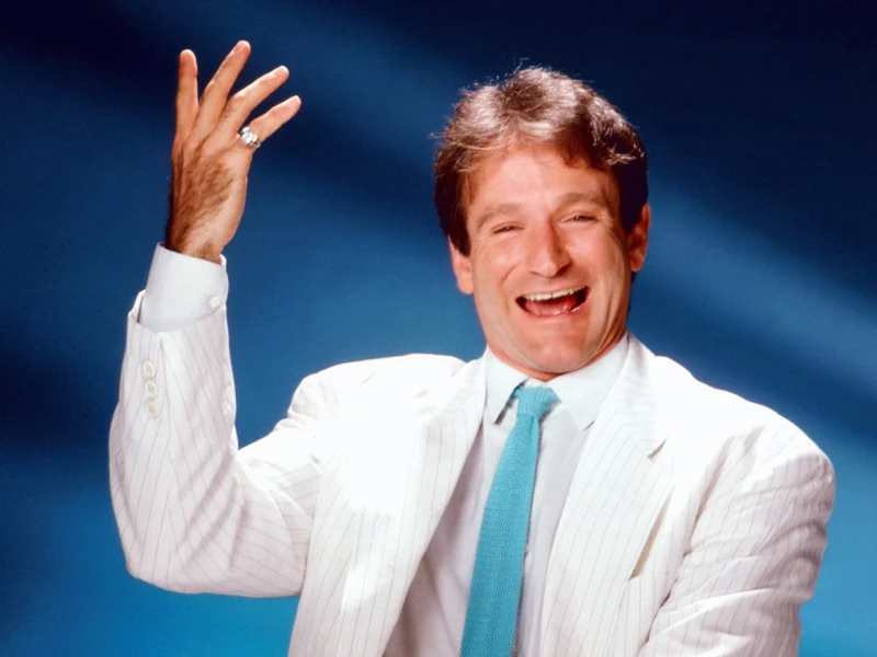 Un joyeux Robin Williams en costume blanc et cravate bleue fait des gestes animés sur un fond bleu. Il semble être en train de rire, levant la main droite dans une pose dynamique.