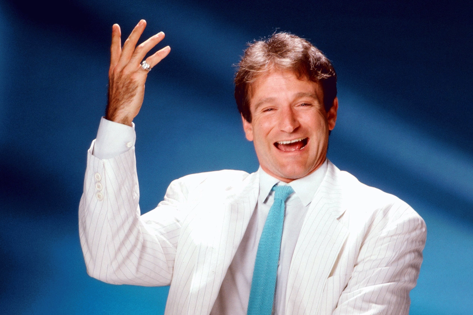 Un joyeux Robin Williams en costume blanc et cravate bleue fait des gestes animés sur un fond bleu. Il semble être en train de rire, levant la main droite dans une pose dynamique.