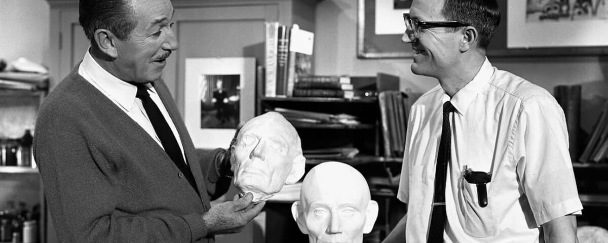 Deux hommes dans un atelier, l'un tenant une tête sculptée de Blaine Gibson et en discutant avec l'autre, entourés d'outils d'art et de sculpture.
