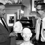 Deux hommes dans un atelier, l'un tenant une tête sculptée de Blaine Gibson et en discutant avec l'autre, entourés d'outils d'art et de sculpture.