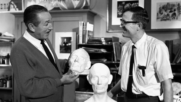 Deux hommes dans un atelier, l'un tenant une tête sculptée de Blaine Gibson et en discutant avec l'autre, entourés d'outils d'art et de sculpture.