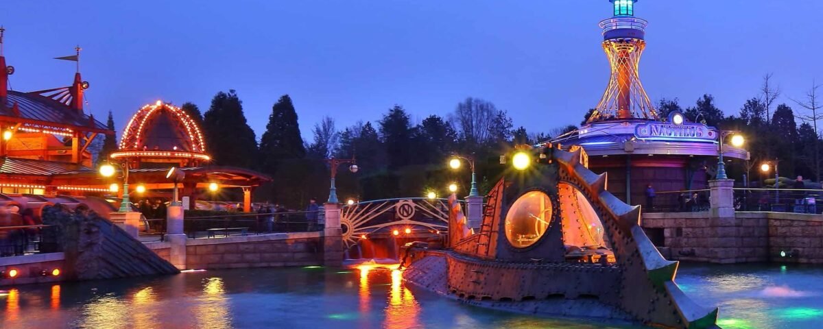 Un parc d'attractions au crépuscule, comprenant un carrousel animé et illuminé, une tour éclairée de couleurs vives et un manège sous-marin "Les Mystères du Nautilus" entouré d'eau éclairée par des néons.