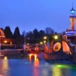 Un parc d'attractions au crépuscule, comprenant un carrousel animé et illuminé, une tour éclairée de couleurs vives et un manège sous-marin "Les Mystères du Nautilus" entouré d'eau éclairée par des néons.