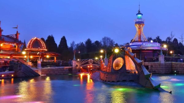 Un parc d'attractions au crépuscule, comprenant un carrousel animé et illuminé, une tour éclairée de couleurs vives et un manège sous-marin "Les Mystères du Nautilus" entouré d'eau éclairée par des néons.