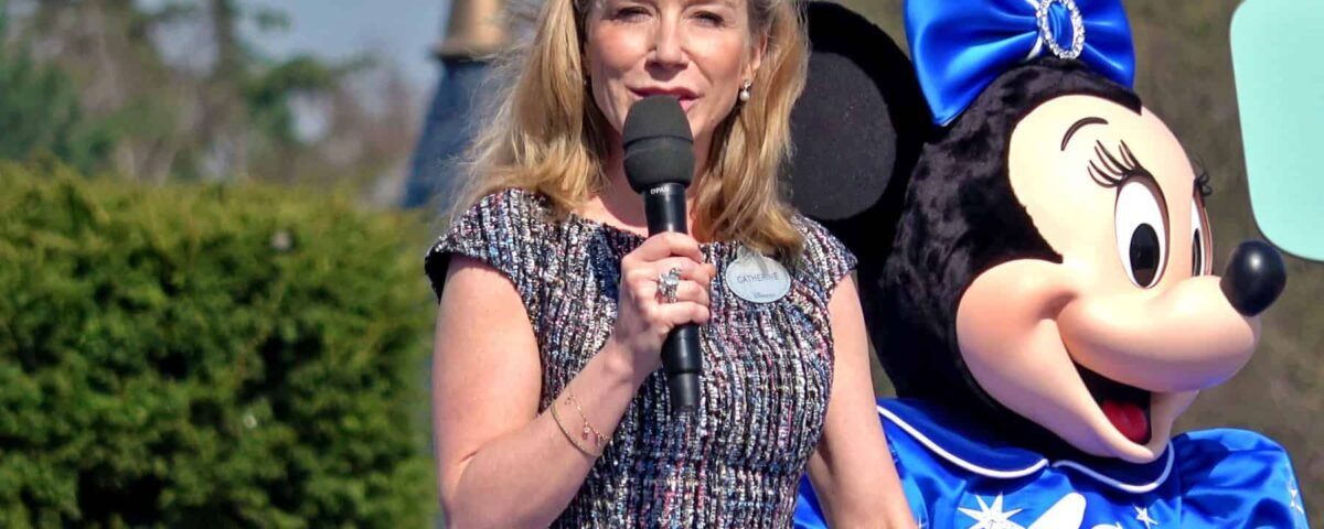 Une femme parle dans un microphone debout à côté de Catherine Powell dans un costume de Minnie Mouse lors d'un événement en plein air, avec un soleil éclatant et de la verdure en arrière-plan.