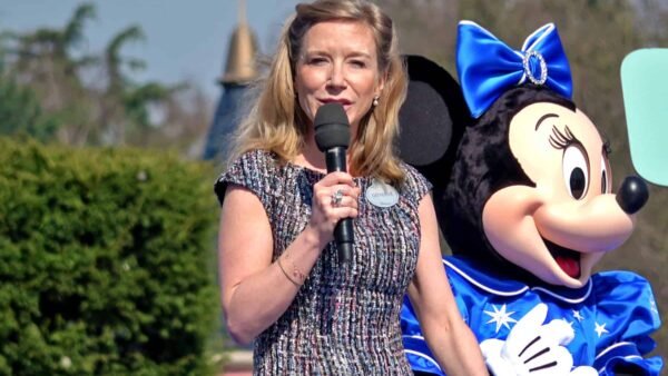 Une femme parle dans un microphone debout à côté de Catherine Powell dans un costume de Minnie Mouse lors d'un événement en plein air, avec un soleil éclatant et de la verdure en arrière-plan.