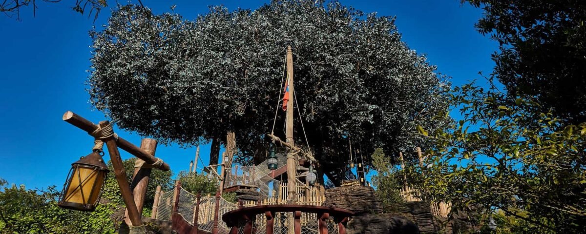 Une cabane dans les arbres robuste, qui rappelle la Cabane des Robinsons de Disneyland, est construite autour d'un grand arbre, dotée de plates-formes en bois et d'une échelle de corde, sur un ciel bleu clair. Une lanterne rustique