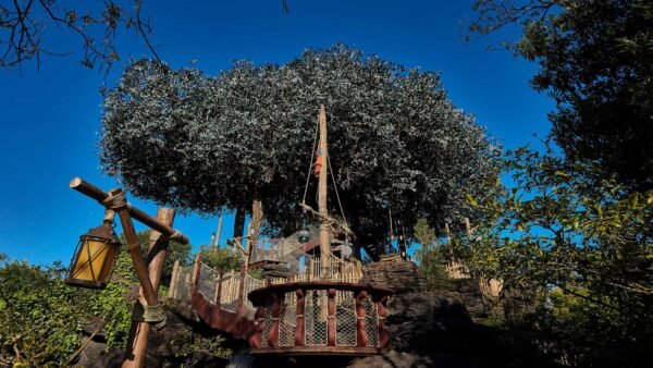 Une cabane dans les arbres robuste, qui rappelle la Cabane des Robinsons de Disneyland, est construite autour d'un grand arbre, dotée de plates-formes en bois et d'une échelle de corde, sur un ciel bleu clair. Une lanterne rustique