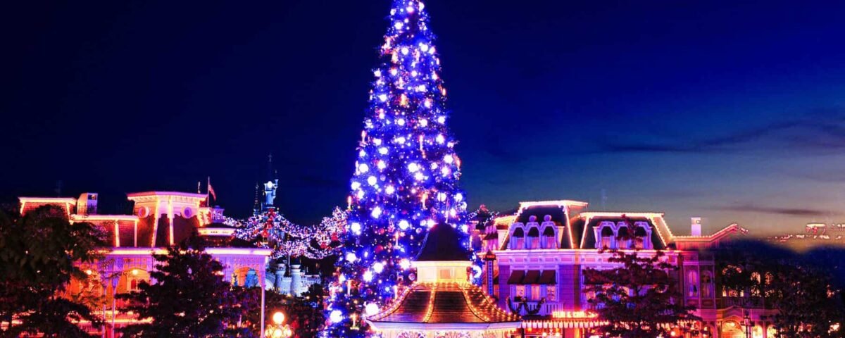 Une scène nocturne animée d'un espace extérieur festif avec un grand sapin bien éclairé, entouré de lumières bleues chatoyantes et de bâtiments décorés.