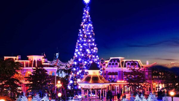 Une scène nocturne animée d'un espace extérieur festif avec un grand sapin bien éclairé, entouré de lumières bleues chatoyantes et de bâtiments décorés.