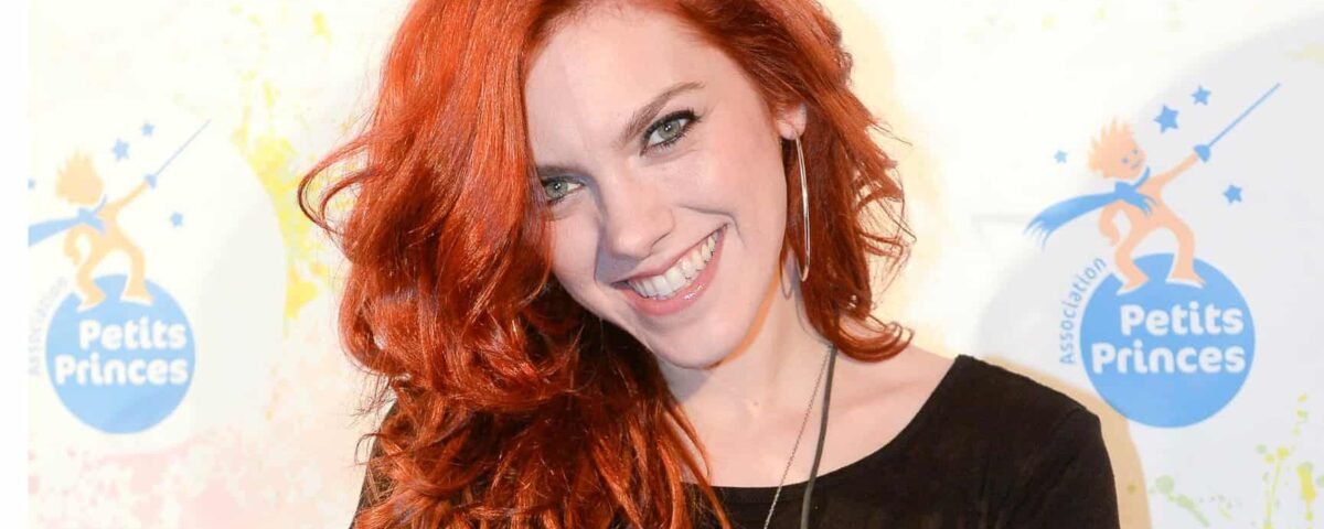 Une femme aux longs cheveux roux bouclés et à la peau claire sourit vivement en posant lors d'un événement. Anaïs Delva porte un haut noir et des boucles d'oreilles, sur fond de logo coloré.