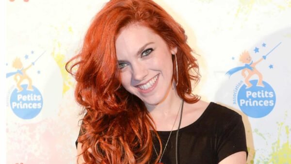 Une femme aux longs cheveux roux bouclés et à la peau claire sourit vivement en posant lors d'un événement. Anaïs Delva porte un haut noir et des boucles d'oreilles, sur fond de logo coloré.