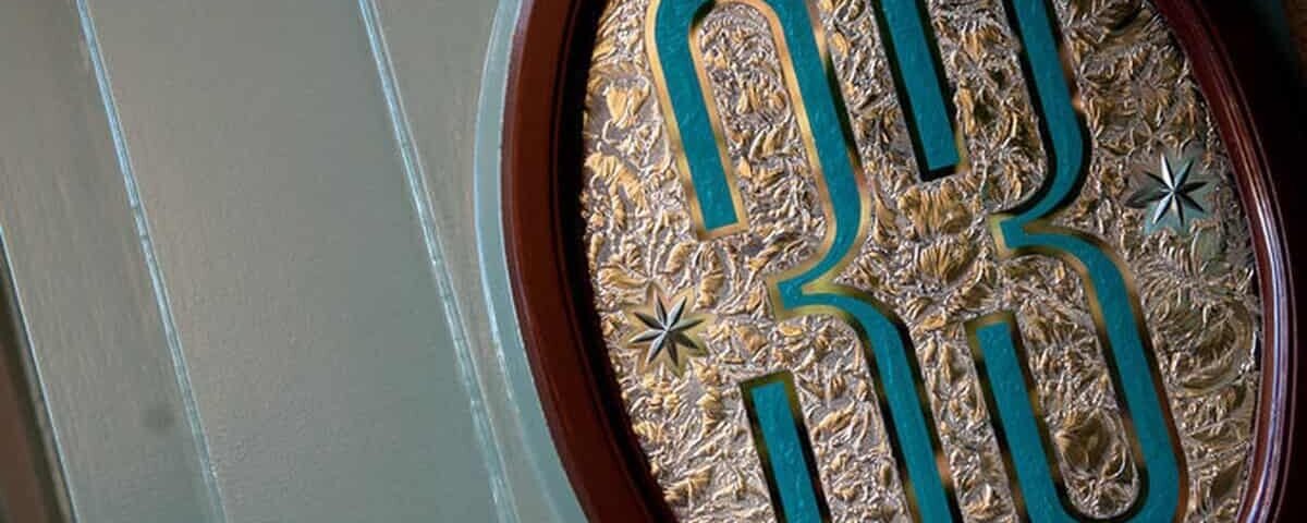 Une plaque décorative avec le numéro « 33 » en relief dans un motif bleu sarcelle et or, sur un fond doré texturé, montée sur une porte en bois vert pâle du Club 33.