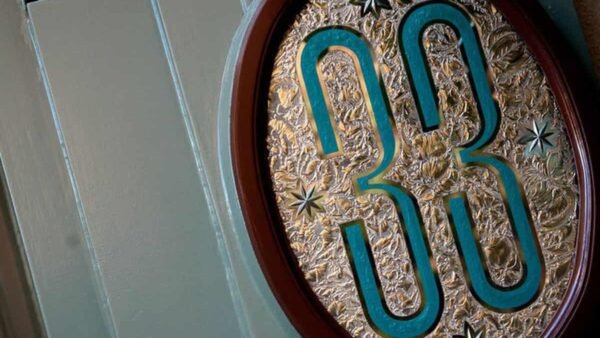 Une plaque décorative avec le numéro « 33 » en relief dans un motif bleu sarcelle et or, sur un fond doré texturé, montée sur une porte en bois vert pâle du Club 33.