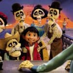 Une scène joyeuse du film Pixar "Coco" mettant en vedette Miguel, entouré de ses ancêtres squelettiques, souriant dans un pays de morts coloré, avec un pont et des pétales de souci brillants au milieu.