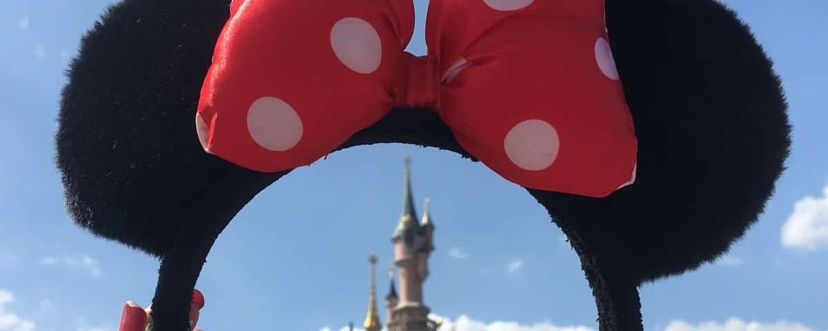 Une paire d'oreilles de Mickey Mouse avec un nœud à pois rouges et blancs, dressées sur un ciel dégagé avec le château de Disneyland Paris en arrière-plan.