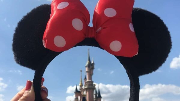 Une paire d'oreilles de Mickey Mouse avec un nœud à pois rouges et blancs, dressées sur un ciel dégagé avec le château de Disneyland Paris en arrière-plan.