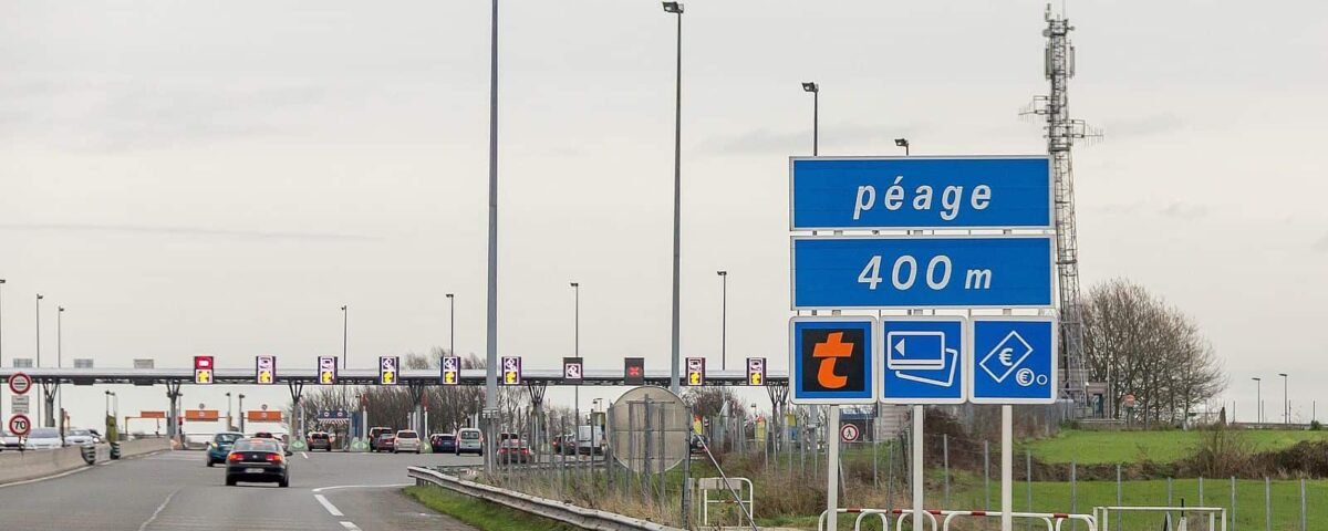 Une autoroute très fréquentée avec un panneau de péage en approche indiquant « péage 400 m » ainsi que des symboles de paiement. Des voitures sont visibles se dirigeant vers les barrières de péage sous un ciel couvert, sur elles