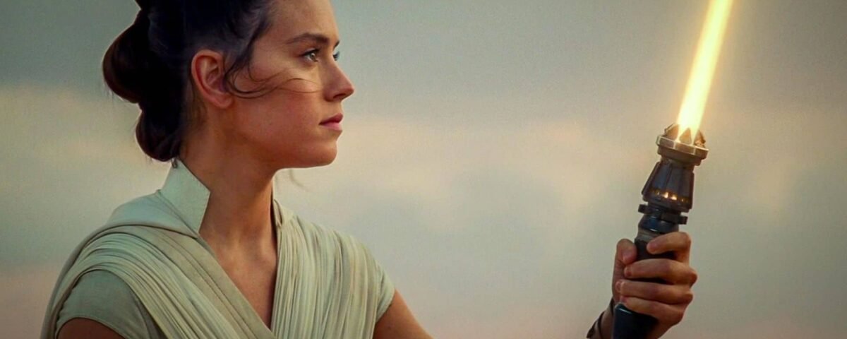 Rey tient un sabre laser vers le ciel au coucher du soleil, son expression étant déterminée et concentrée, dans un paysage désertique serein.