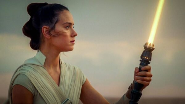 Rey tient un sabre laser vers le ciel au coucher du soleil, son expression étant déterminée et concentrée, dans un paysage désertique serein.