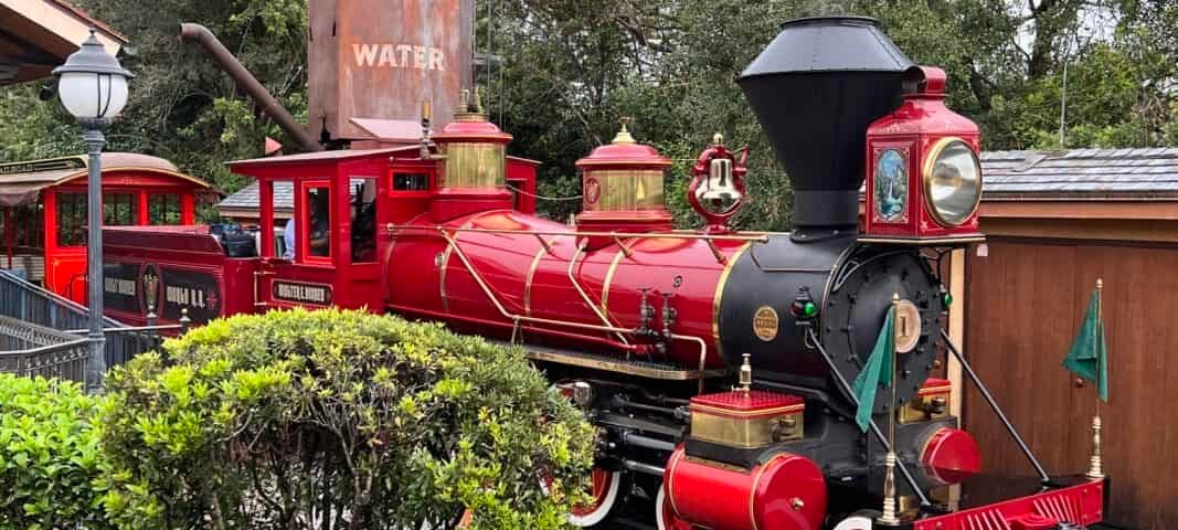 Une locomotive vintage rouge vif avec un drapeau vert se dresse sur des voies entourées d'une verdure luxuriante et d'un réservoir « d'eau » en arrière-plan sous un ciel nuageux.