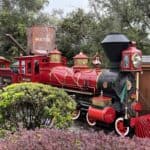 Une locomotive vintage rouge vif avec un drapeau vert se dresse sur des voies entourées d'une verdure luxuriante et d'un réservoir « d'eau » en arrière-plan sous un ciel nuageux.