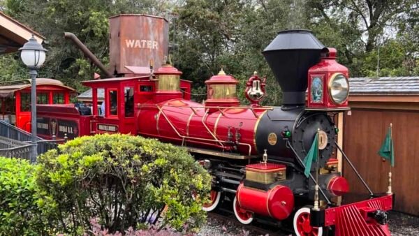 Une locomotive vintage rouge vif avec un drapeau vert se dresse sur des voies entourées d'une verdure luxuriante et d'un réservoir « d'eau » en arrière-plan sous un ciel nuageux.