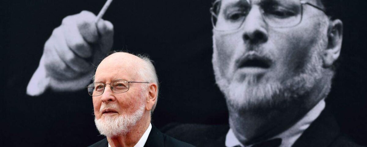 Un homme âgé en smoking assiste à un événement Star Wars, debout devant une grande image monochrome de John Williams en action.
