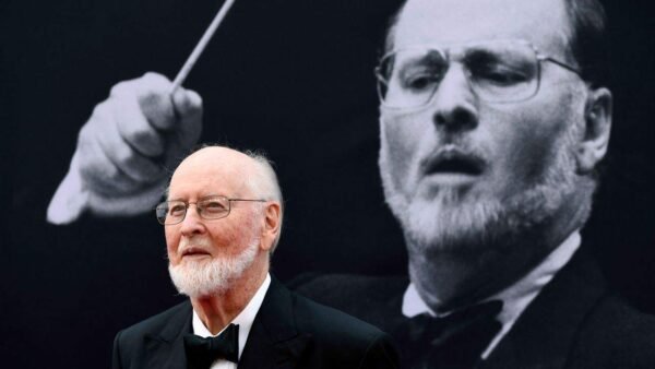 Un homme âgé en smoking assiste à un événement Star Wars, debout devant une grande image monochrome de John Williams en action.