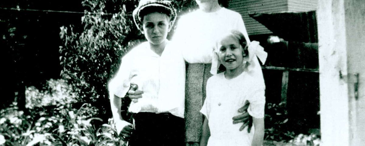Une photo vintage en noir et blanc d'une femme debout avec son bras autour d'un jeune garçon qui tient un petit oiseau, et d'une jeune fille debout à côté d'eux, le tout devant une flore et une maison.
