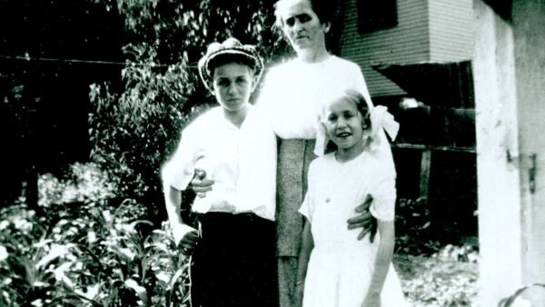 Une photo vintage en noir et blanc d'une femme debout avec son bras autour d'un jeune garçon qui tient un petit oiseau, et d'une jeune fille debout à côté d'eux, le tout devant une flore et une maison.