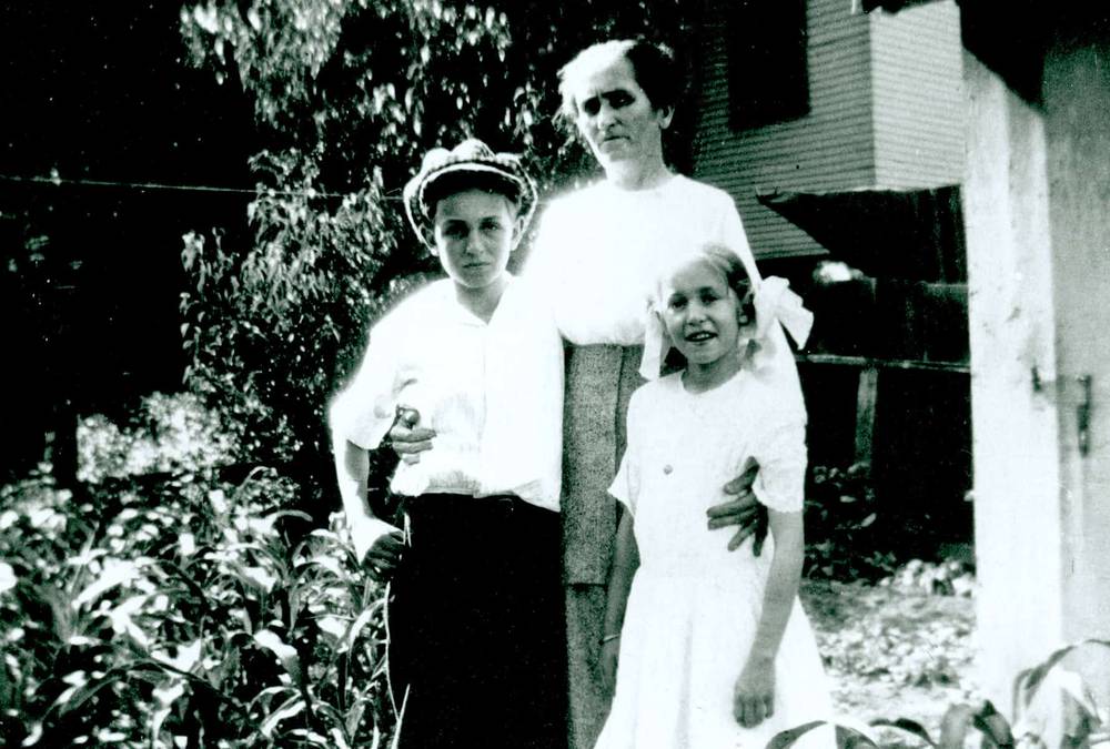 Une photo vintage en noir et blanc d'une femme debout avec son bras autour d'un jeune garçon qui tient un petit oiseau, et d'une jeune fille debout à côté d'eux, le tout devant une flore et une maison.