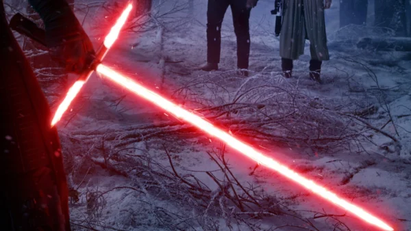 Un laser sabre crossguard rouge brillant se trouve sur un sol couvert de givre dans une forêt enneigée, avec deux personnages sombres debout en arrière-plan.